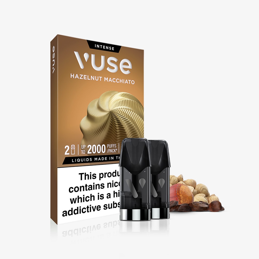 A pack of Vuse Hazelnut Macchiato Extra Intense Flavour Pods, with two coffee-flavoured vape pods in front, a swirl-designed box, mixed nuts including hazelnuts beside them, and a nicotine warning label below.